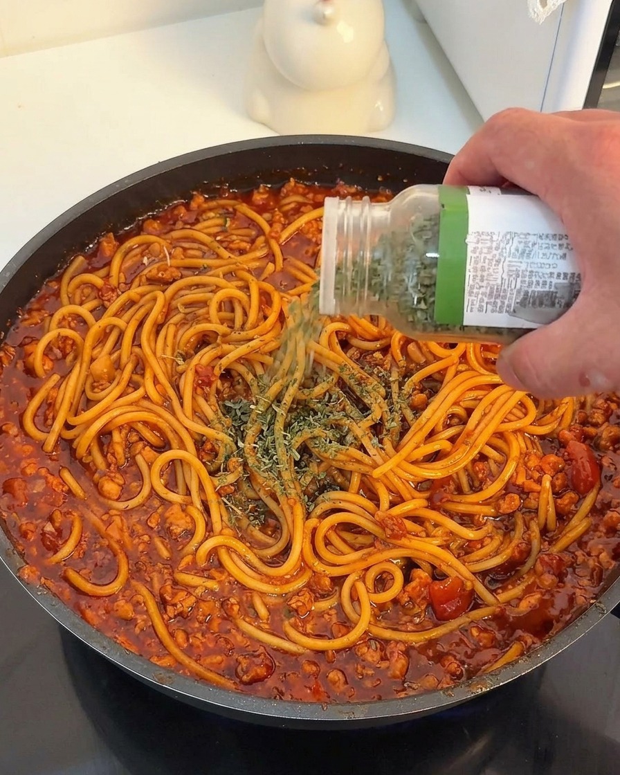 A hand holding a clear spice jar with a green lid, sprinkling dried parsley flakes over spaghetti coated in a rich red tomato meat sauce.