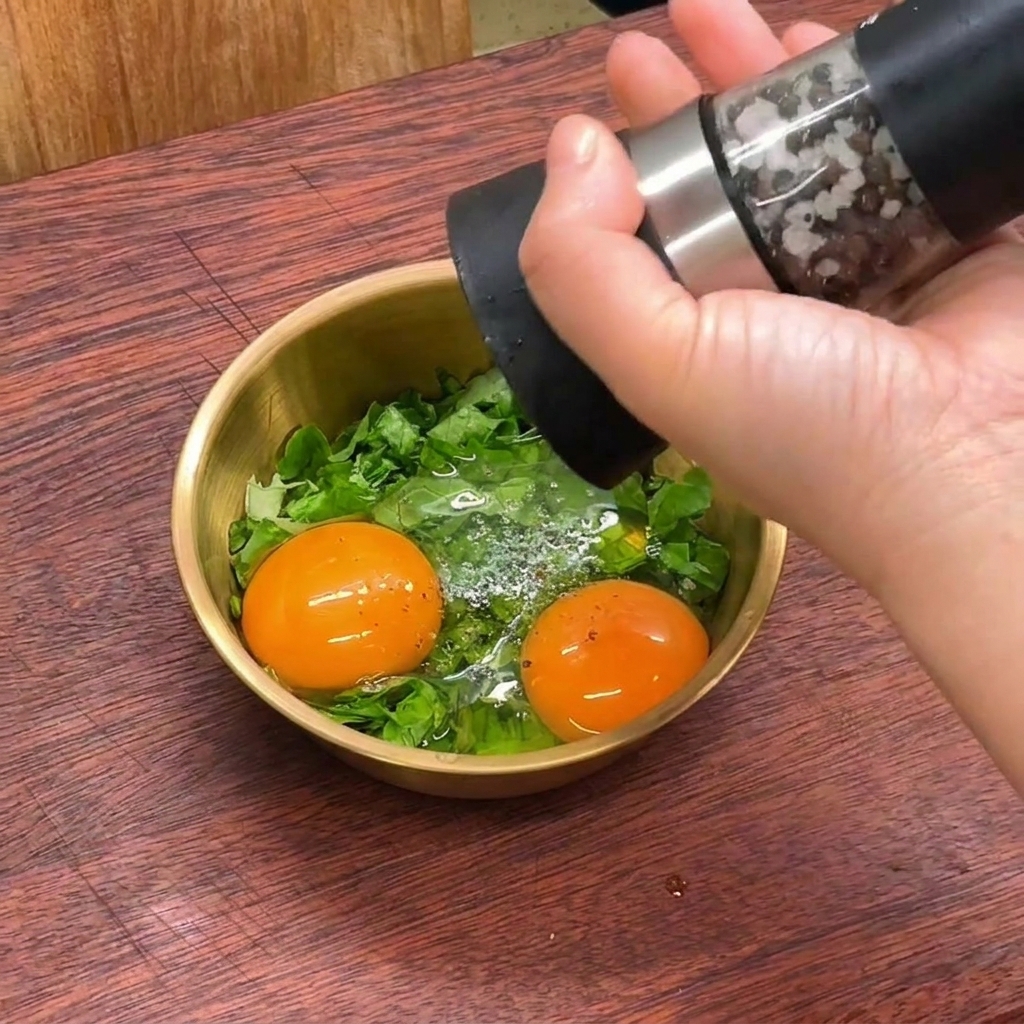 A hand using a pepper mill to grind fresh black pepper into a small gold bowl containing raw eggs and shredded green lettuce.