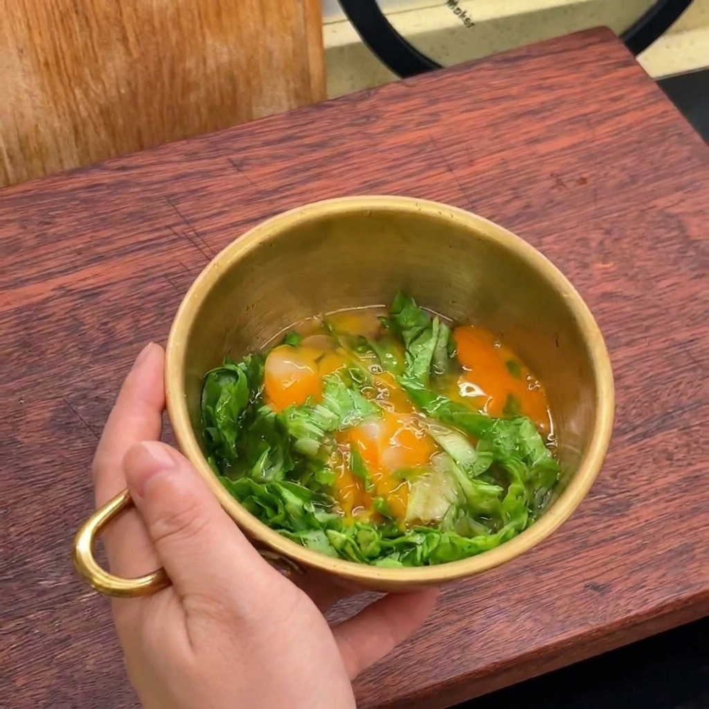 A hand holding a gold bowl filled with chopped green lettuce and two freshly cracked raw eggs.