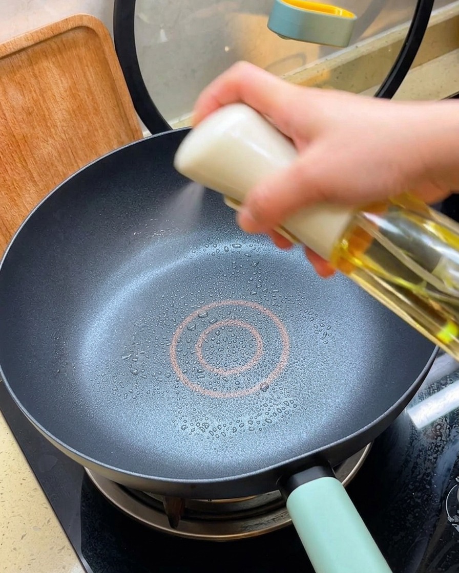 A hand holding an oil spray bottle, misting a thin layer of cooking oil into a heated black non-stick pan.