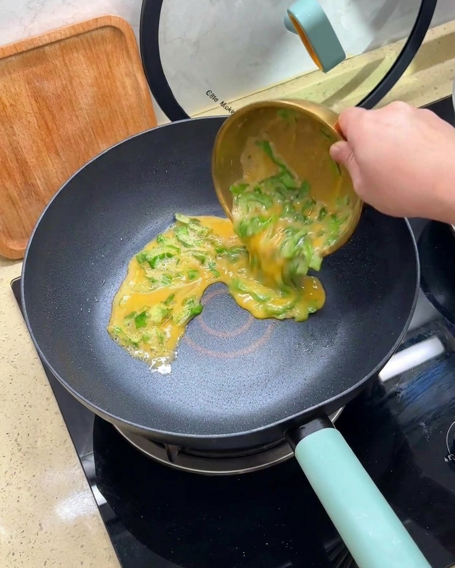Pouring a mixture of beaten eggs and chopped lettuce from a gold bowl into a hot non-stick frying pan.