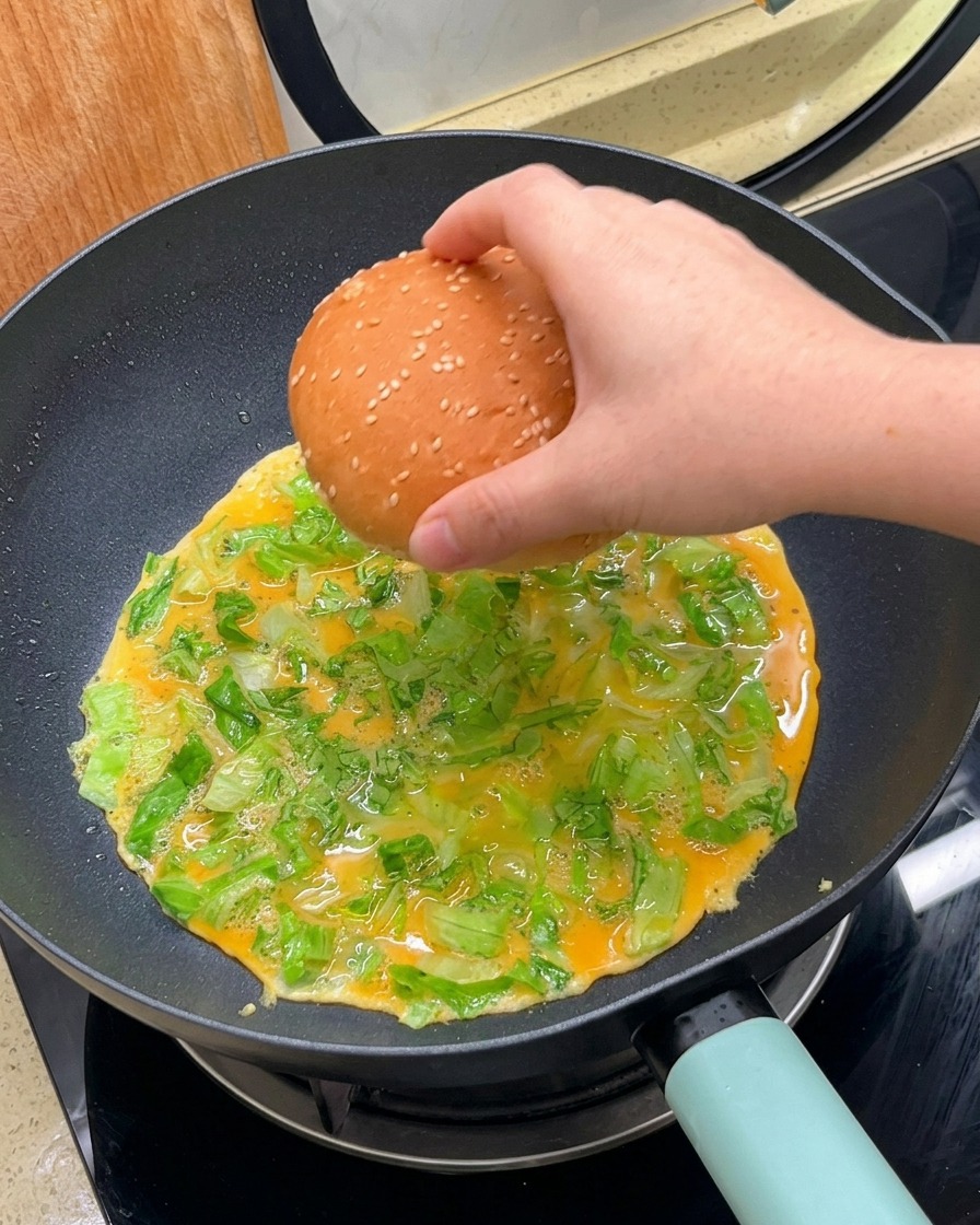 A hand placing a sesame seed burger bun face-down onto a cooking egg and lettuce mixture in a pan.