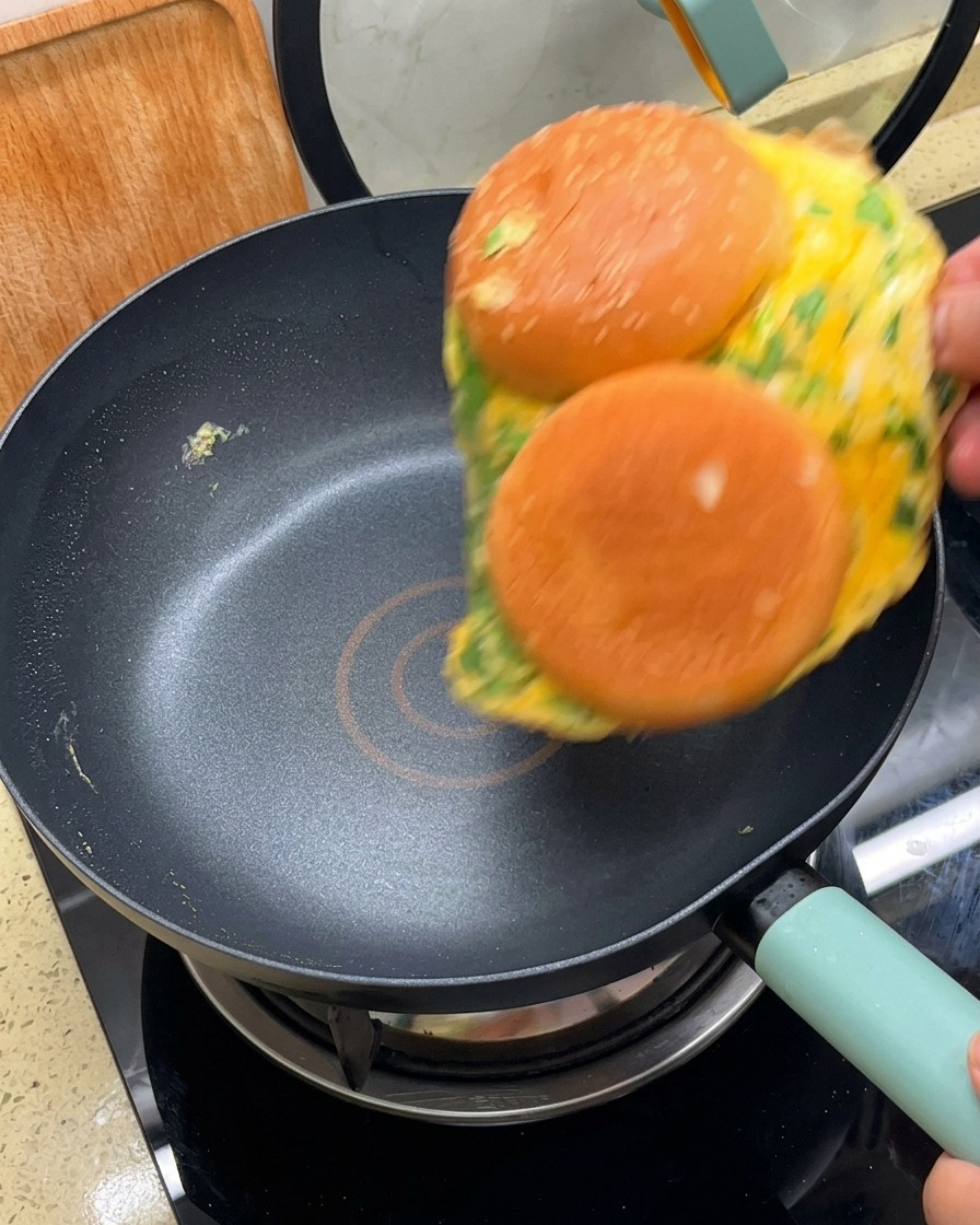 An omelet with two burger bun halves attached being flipped over in a black non-stick skillet.