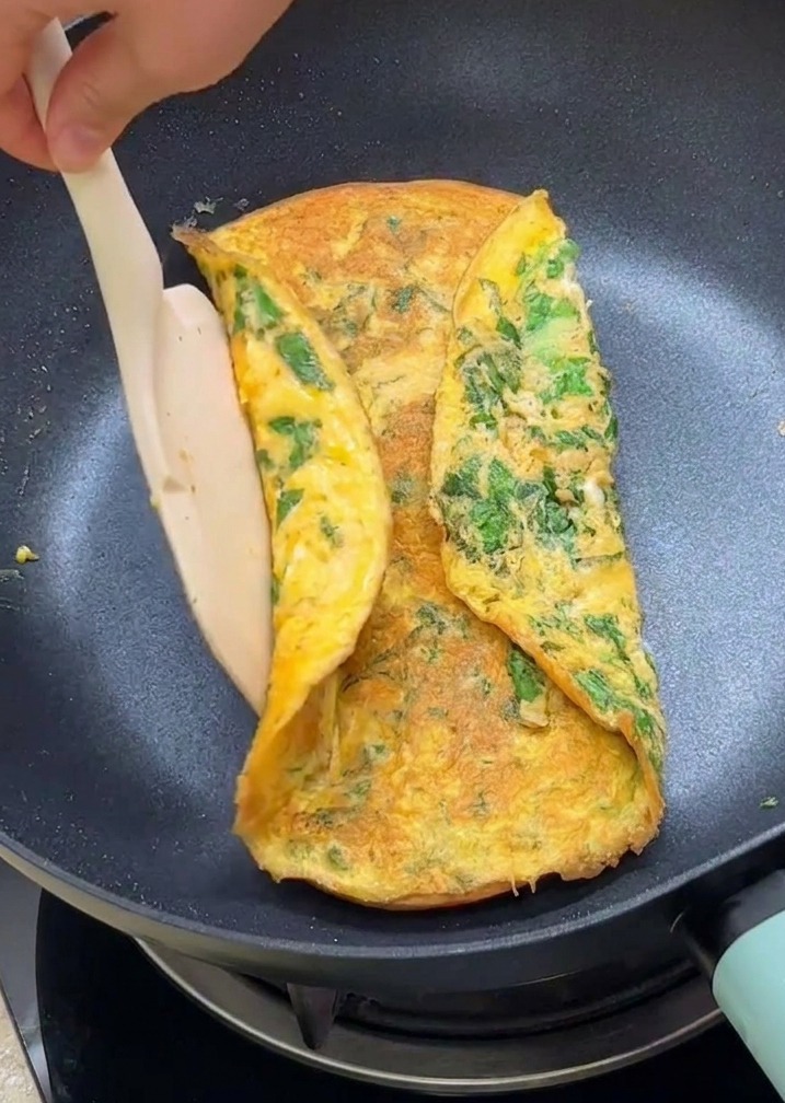A spatula folding the edges of a green lettuce omelet over two toasted burger bun halves in a non stick frying pan.