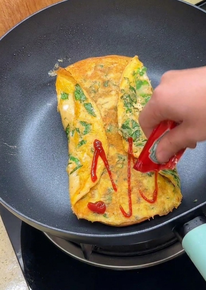 A hand squeezing red ketchup from a packet onto the folded omelet surface inside a pan.