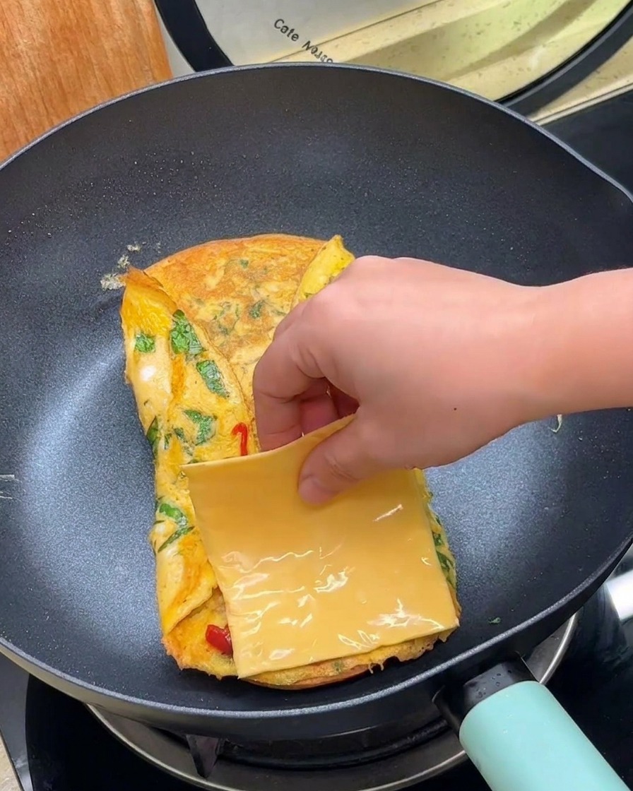 A hand placing a square slice of yellow cheese onto a folded egg inside a frying pan.