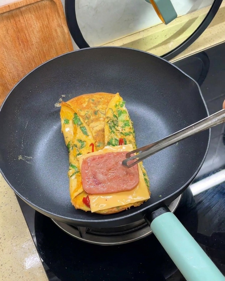 Tongs placing a slice of cooked meat over a slice of cheese on a folded omelet in a pan.