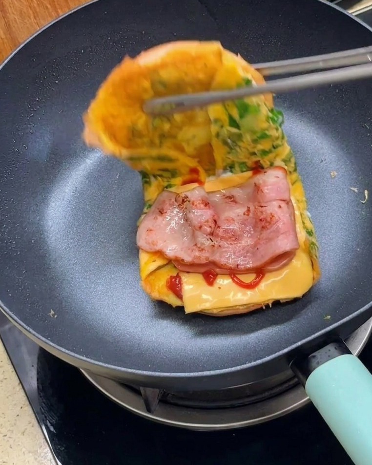 Tongs folding the two halves of the omelet-wrapped burger bun together to form a finished sandwich.