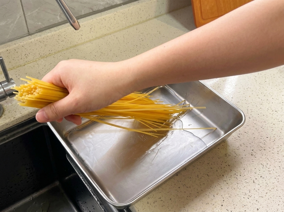 A hand placing a bundle of dry yellow spaghetti into a shallow rectangular metal tray filled with water on a kitchen counter.