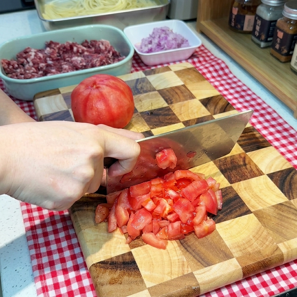 A hand using a wide knife to dice a peeled red tomato into small cubes on a checkered wooden cutting board.