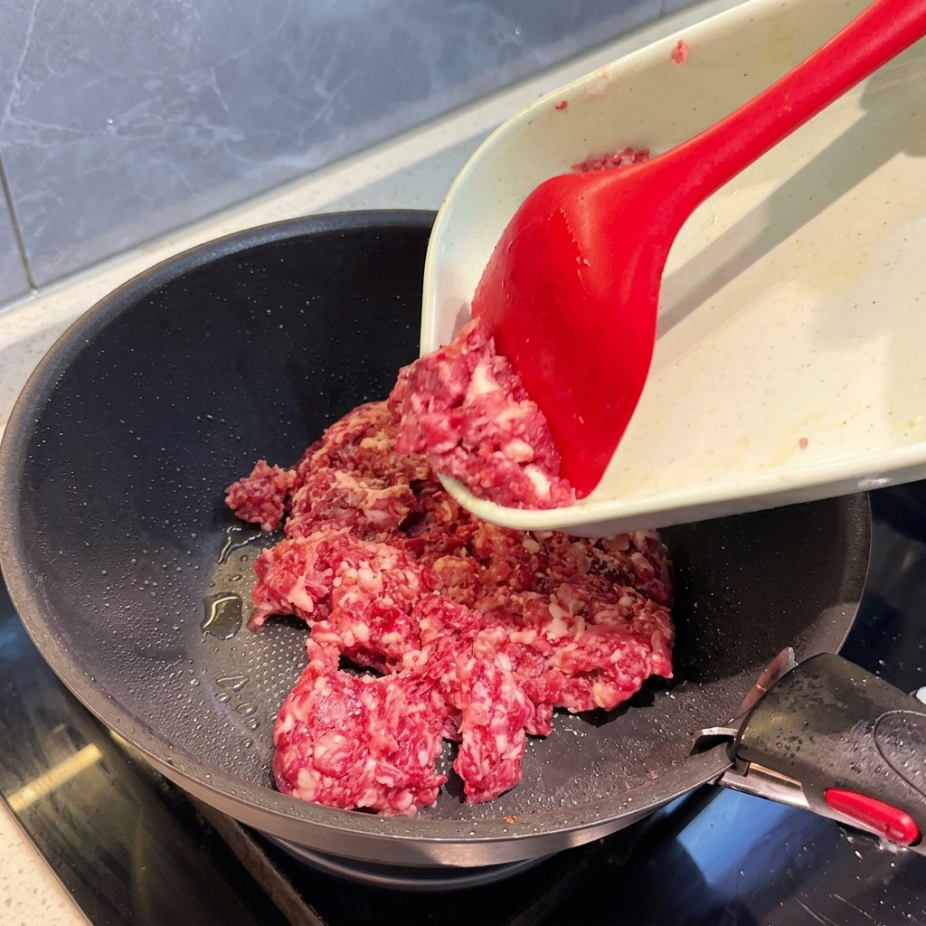 Raw, coarsely ground mixed meat being pushed from a cutting board into a dark skillet using a bright red spatula.