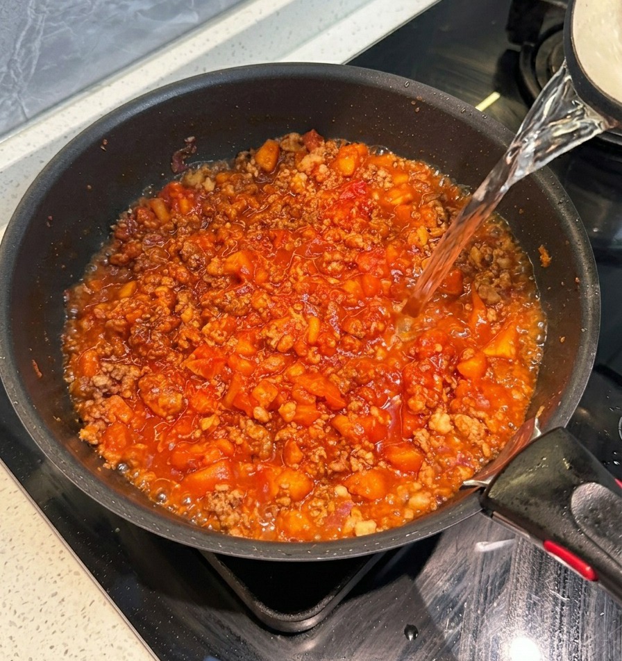 Pouring clear hot water from a kettle into a black skillet containing a thick red tomato meat sauce.