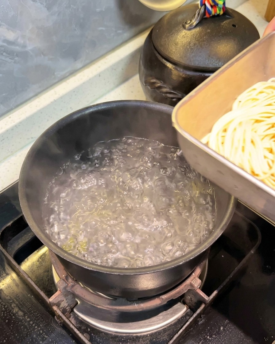 Placing a portion of long spaghetti noodles from a metal tray into a black pot of bubbling boiling water.