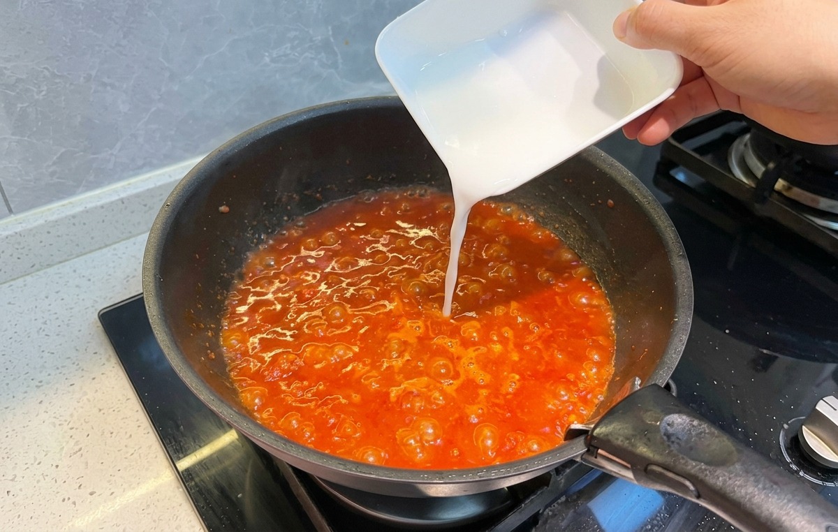 Pouring a milky white starch slurry from a small square dish into a pan of bubbling red tomato meat sauce.