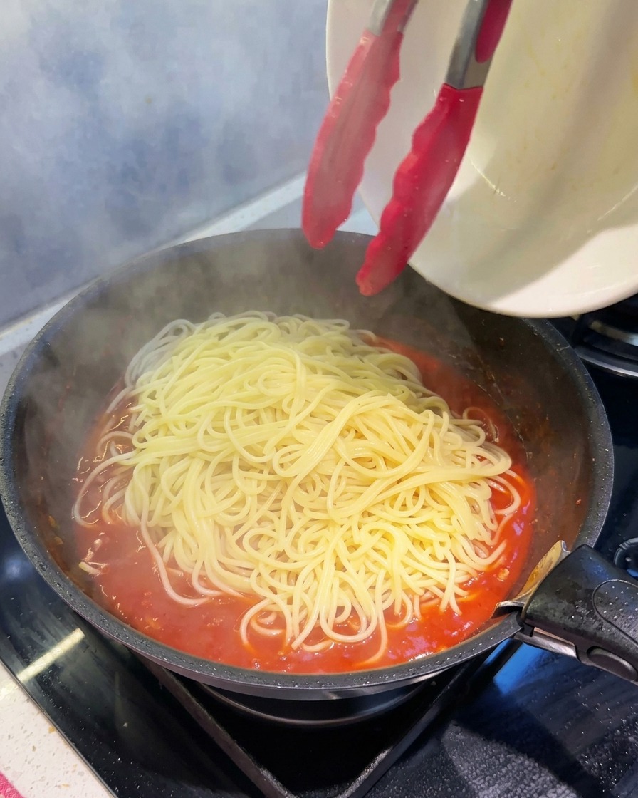 Tongs transferring steaming, freshly cooked spaghetti into a skillet full of thick, bubbling red tomato meat sauce.