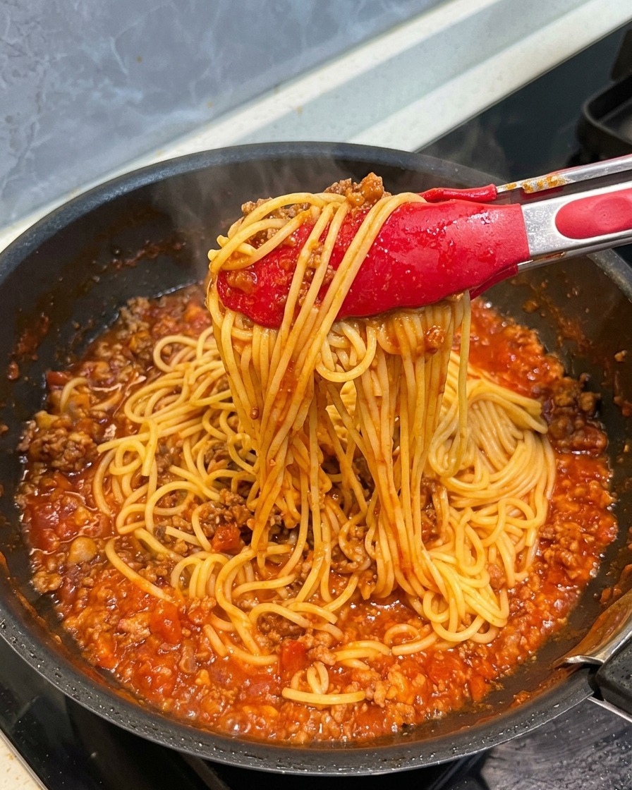 Red tongs pulling up a bunch of spaghetti noodles, showing them thoroughly coated in a rich and hearty tomato meat sauce.