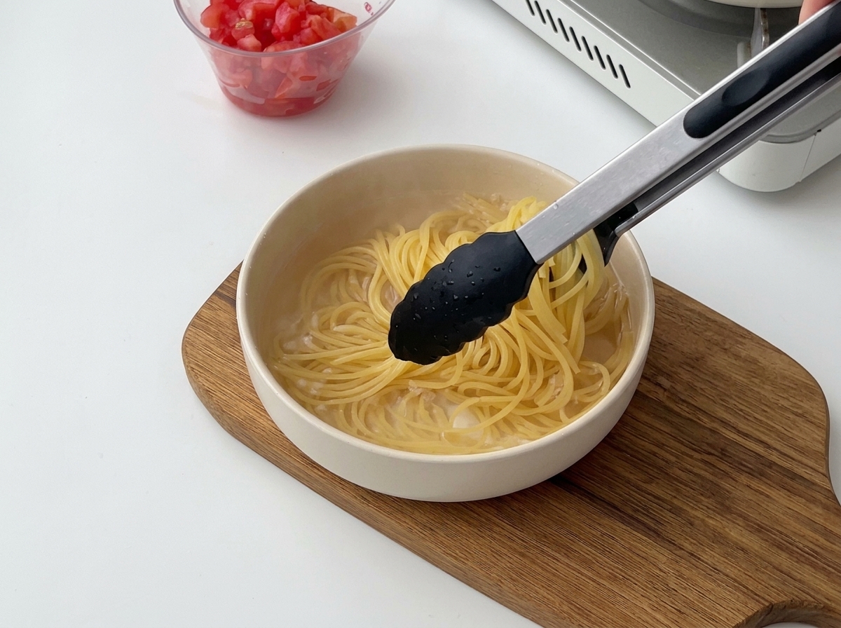 Black tongs lifting cooked spaghetti from hot water and transferring it to a bowl of cold water.