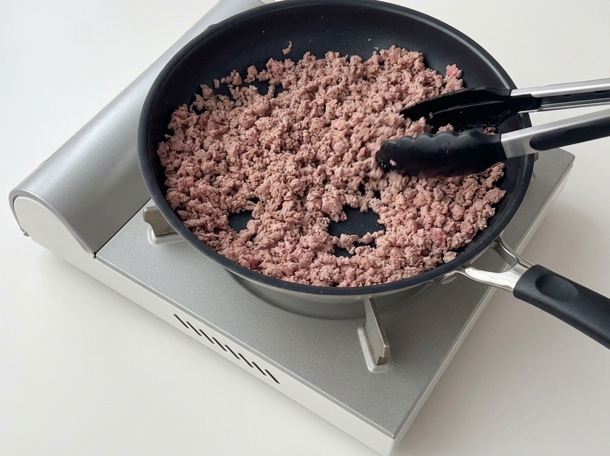 Tongs breaking apart and stirring browned, crumbly minced beef in a non-stick skillet.