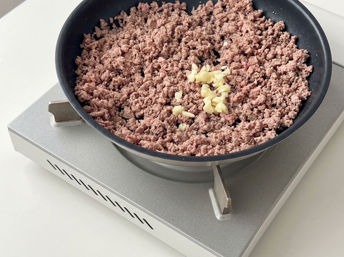 Minced garlic placed on top of browned minced beef in a dark frying pan.