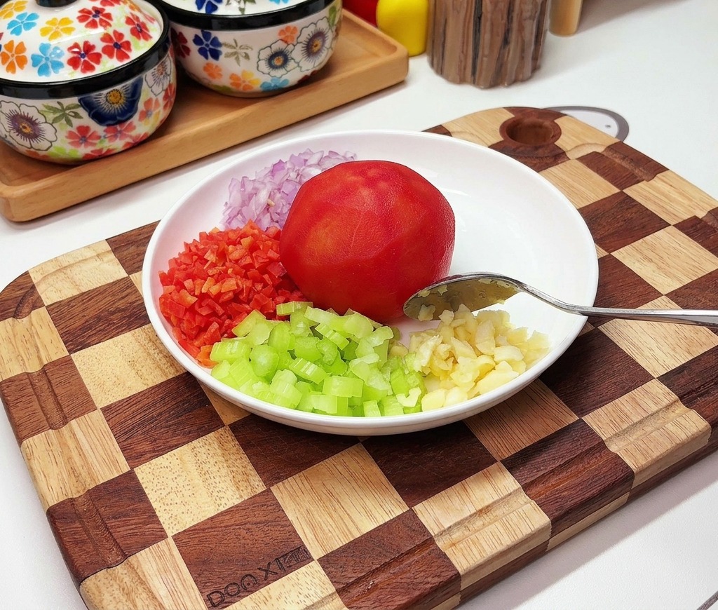 A white plate holding a peeled tomato, surrounded by neatly diced red onion, carrot, celery, and minced garlic.