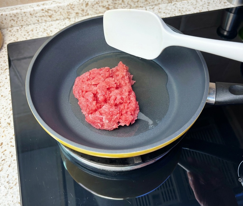A block of raw minced beef being placed into a heated skillet with oil using a white spatula.