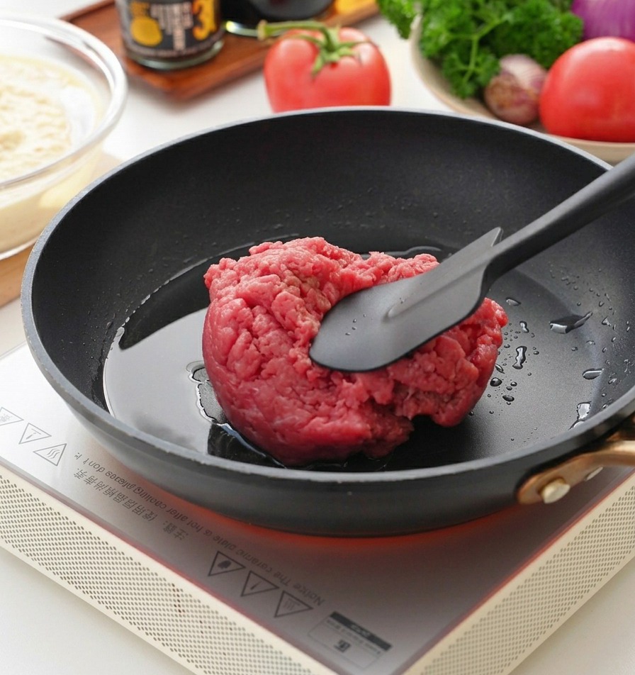 A dark spatula pressing into a mound of raw ground beef in a hot black frying pan.