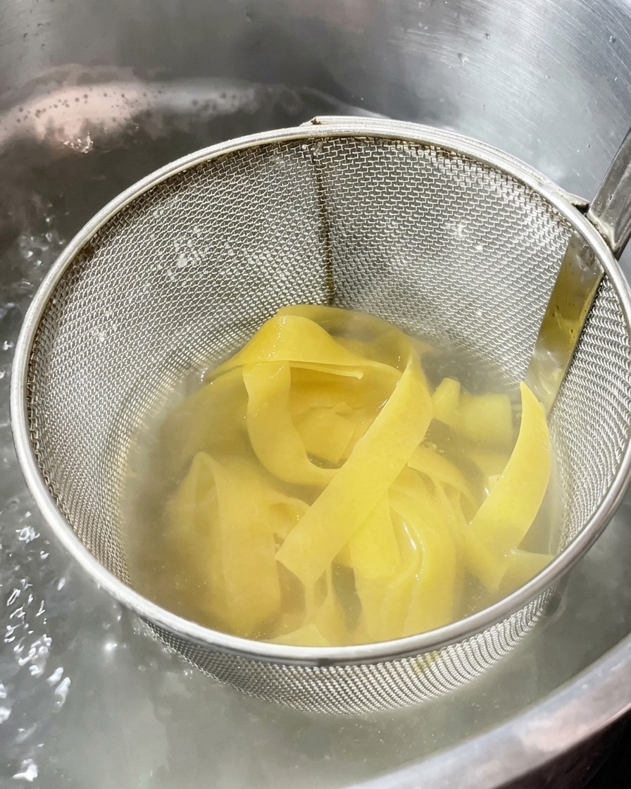 Wide ribbon pasta cooking inside a metal mesh strainer submerged in a pot of boiling water.