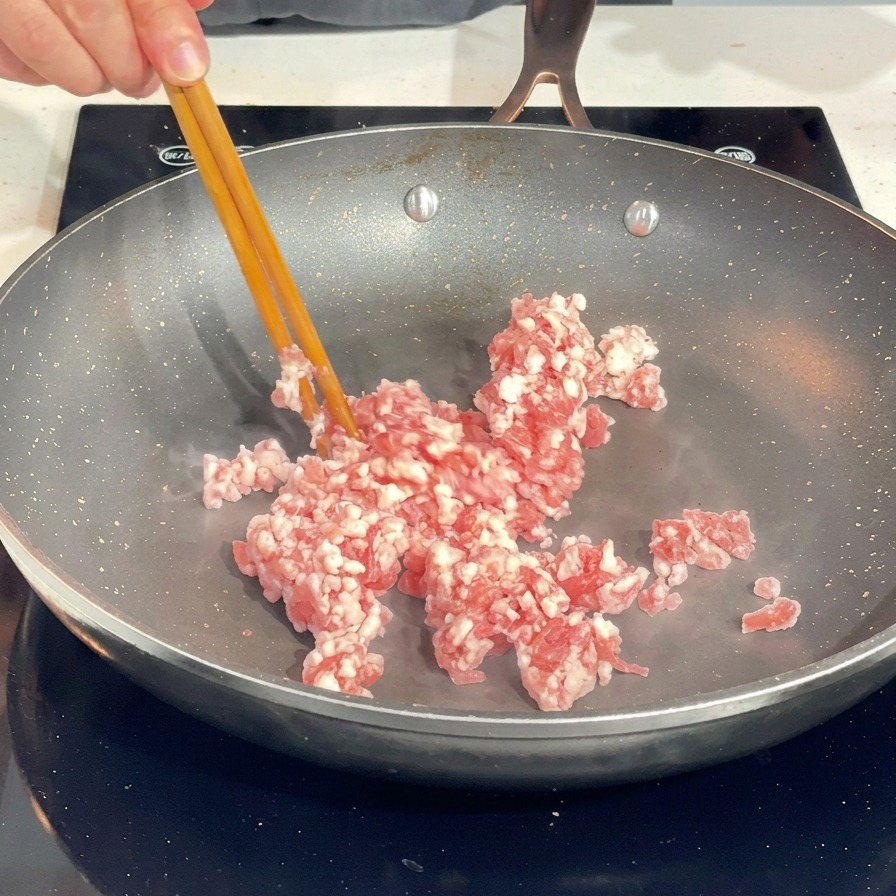 Using chopsticks to break apart raw minced pork in a hot frying pan.