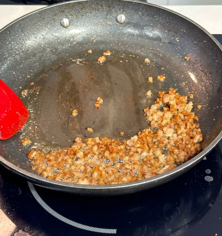 Minced pork browning in a dark non-stick pan, being stirred with a bright red spatula.