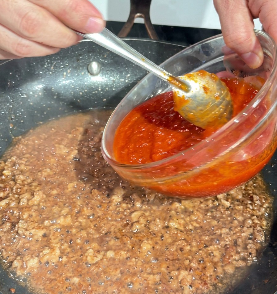 Pouring thick red tomato sauce from a glass bowl into a pan of browned minced pork.