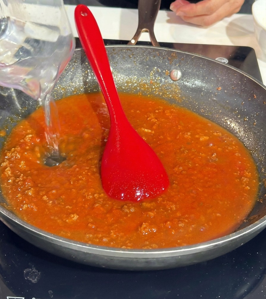Pouring water from a clear measuring cup into a pan of rich red tomato meat sauce.