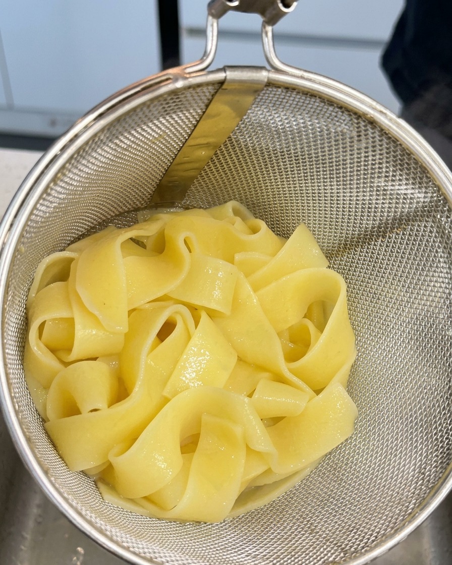 Cooked wide ribbon pasta resting in a metal mesh strainer after being pulled from boiling water.