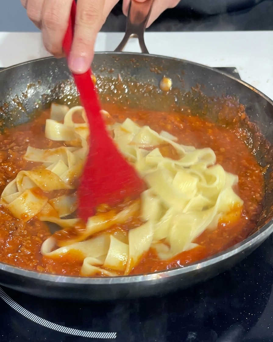 A red spatula tossing thick pasta ribbons in a skillet, coating them evenly with a thick tomato and meat sauce.