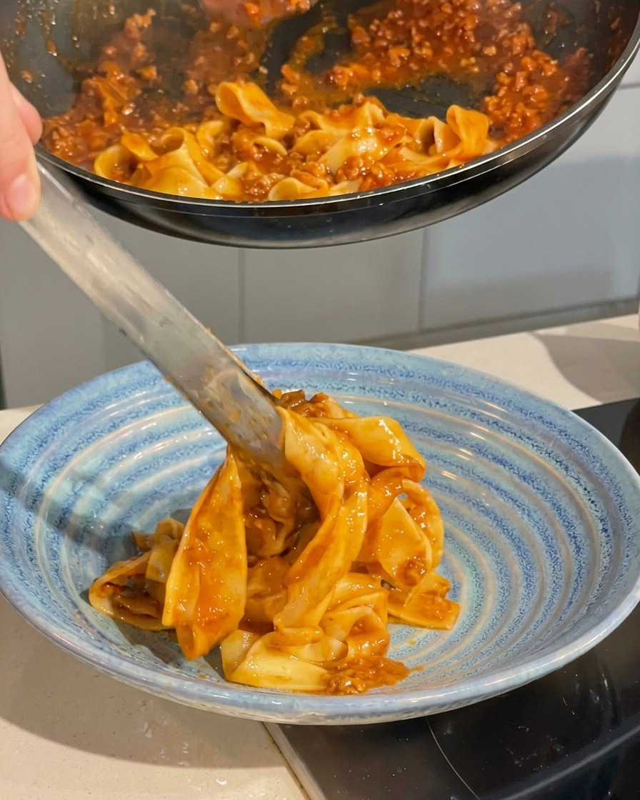 Metal tongs lifting saucy wide pasta ribbons from a black frying pan and plating them into a deep blue ceramic serving bowl.