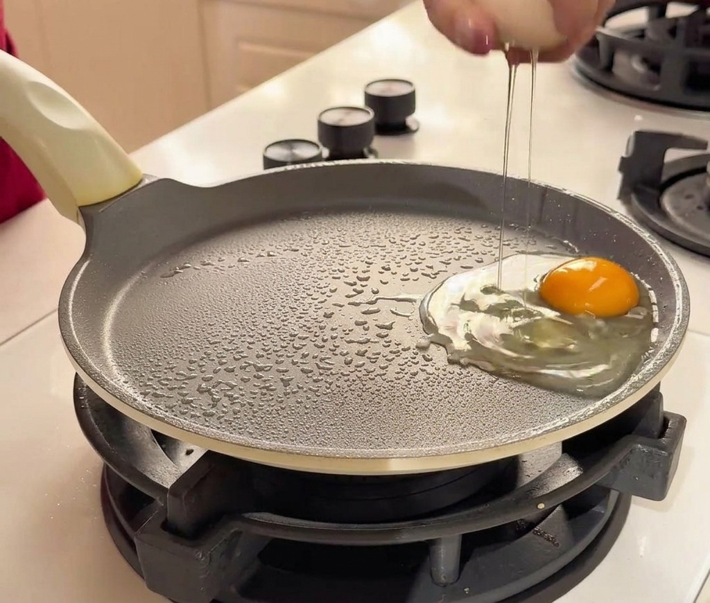 A hand cracking a fresh egg into a non-stick frying pan next to another cracked egg.