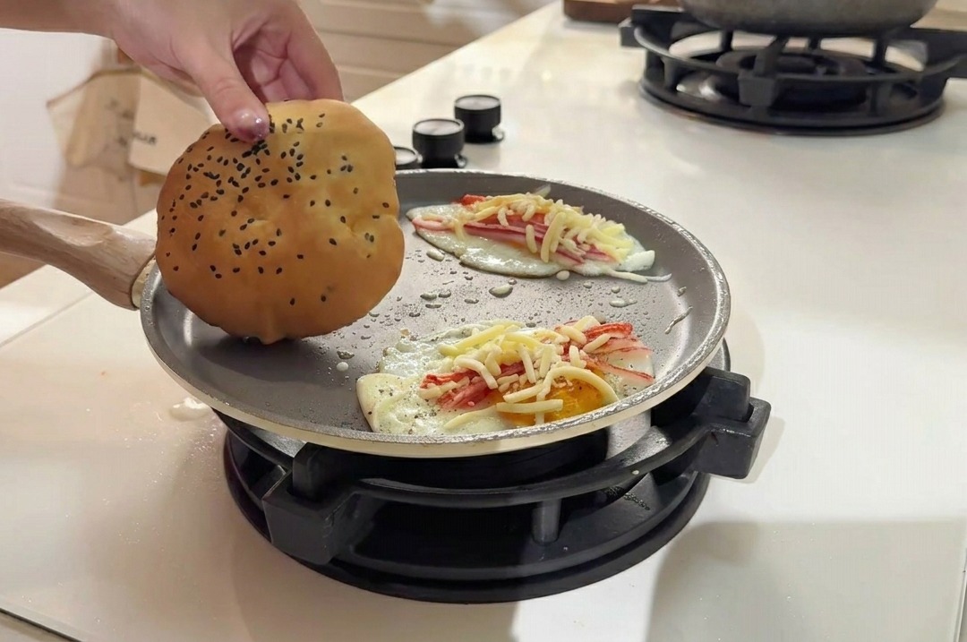 Placing a golden sesame seed burger bun onto the pan surface alongside the cooking egg patties.