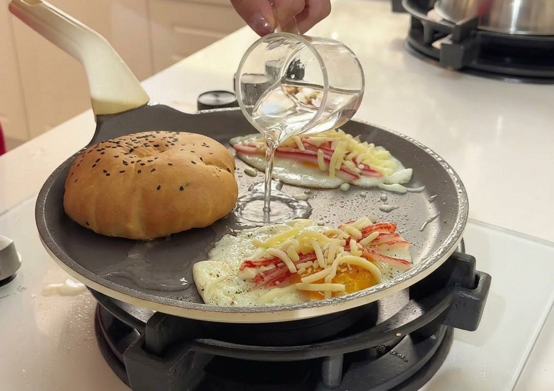 Pouring clear water from a small glass cup into the hot frying pan between the bun and the eggs.