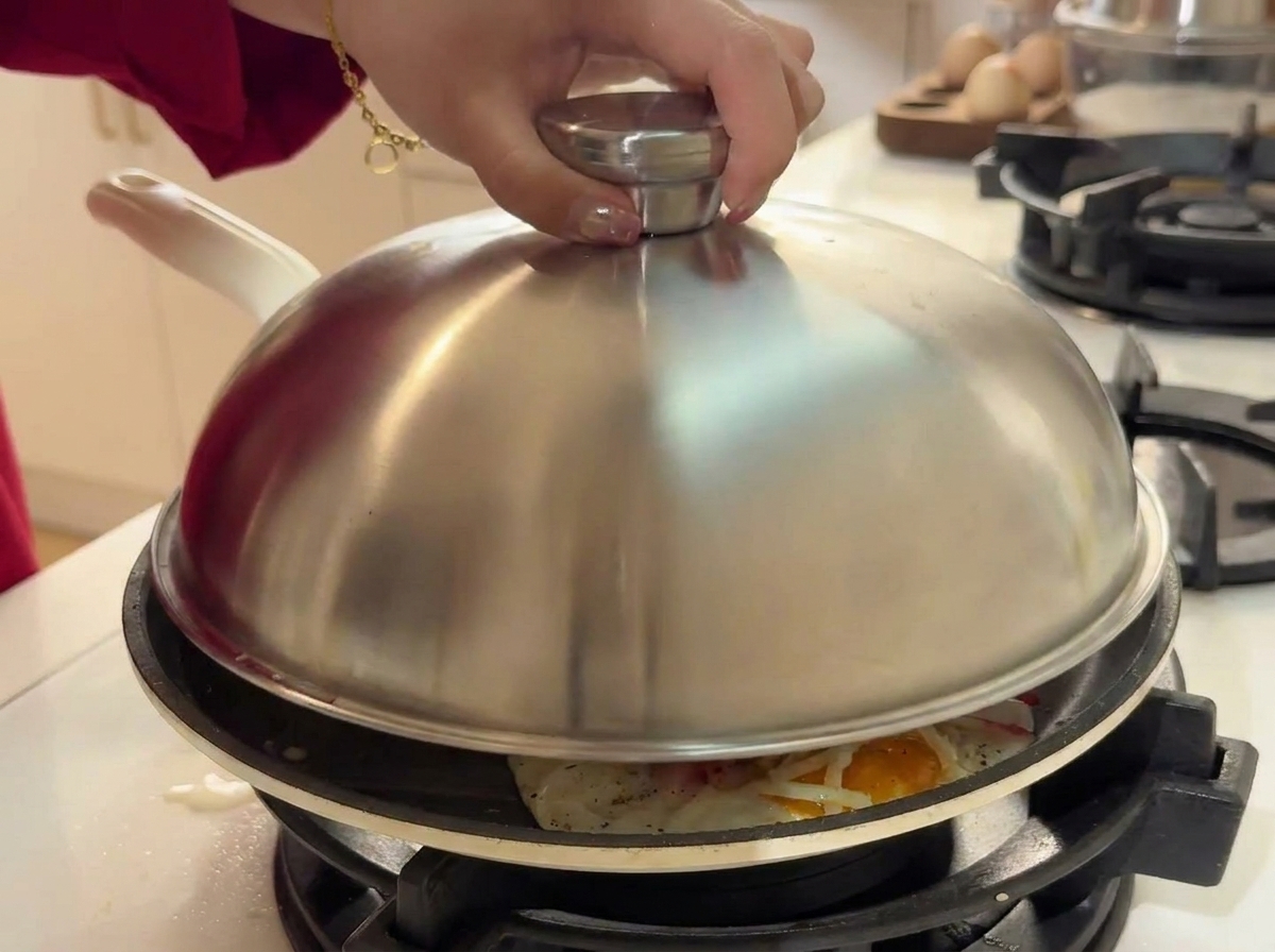 A hand placing a domed stainless steel lid over a skillet containing cooking eggs, shredded cheese, and crab sticks.