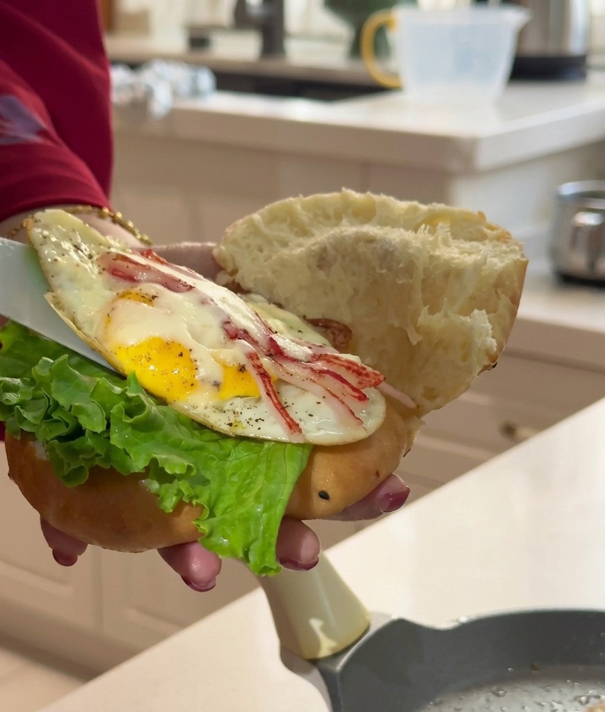A hand holding an open steamed bun with green lettuce, while a white spatula slides a cooked egg and crab patty inside.