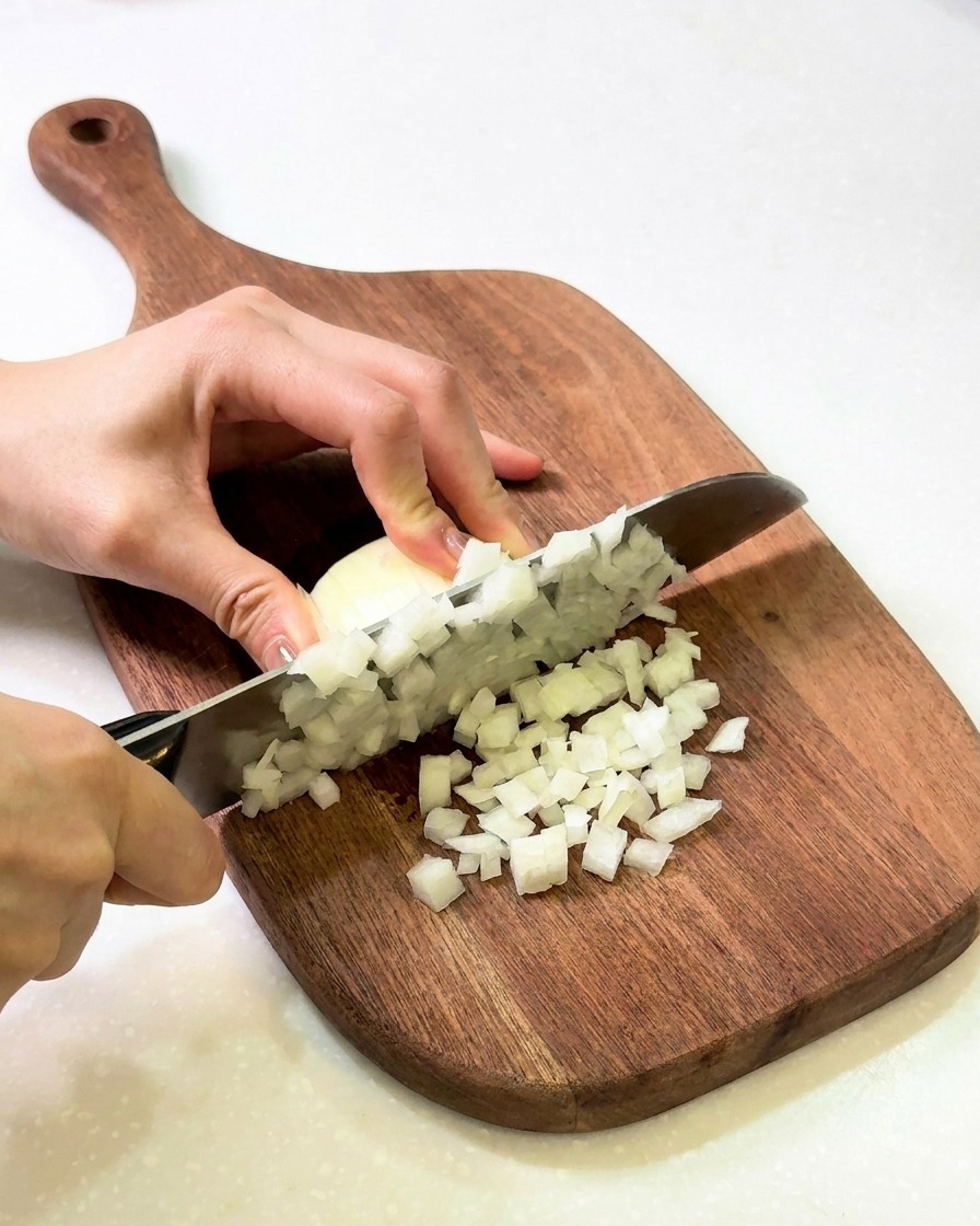 Hands chopping a white onion into fine dice on a wooden cutting board.