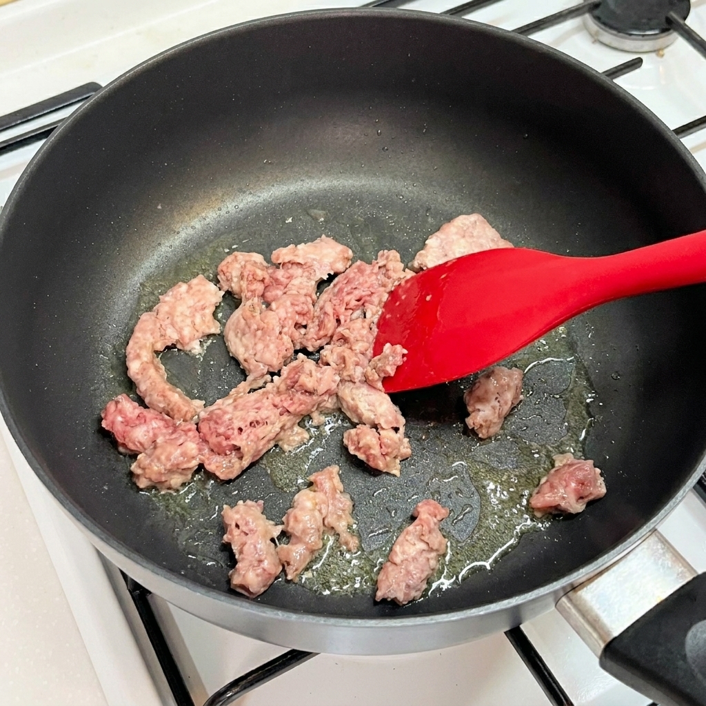 Raw pink minced beef being broken apart and stirred in a hot skillet with a red silicone spatula.