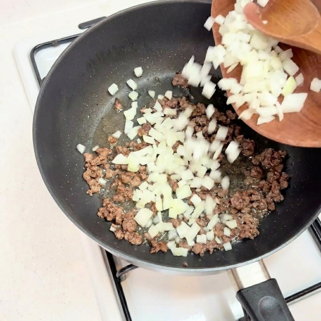 Chopped white onions being added to browned minced beef in a black skillet.