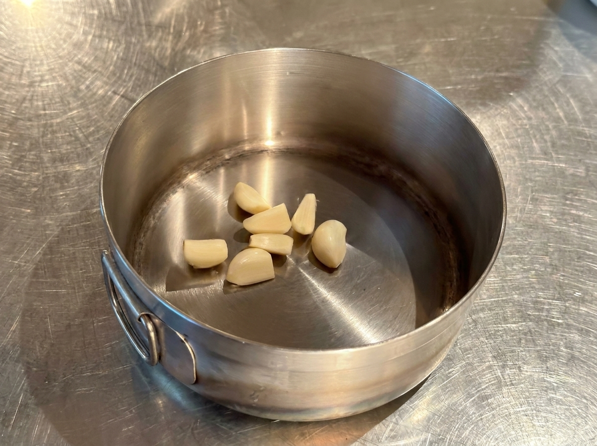 Several whole peeled garlic cloves resting at the bottom of a small, dry stainless steel pot.