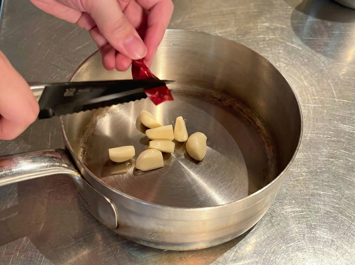 Hands using a small black knife or scissors to cut a dried red chili pepper into pieces over a pot of garlic.