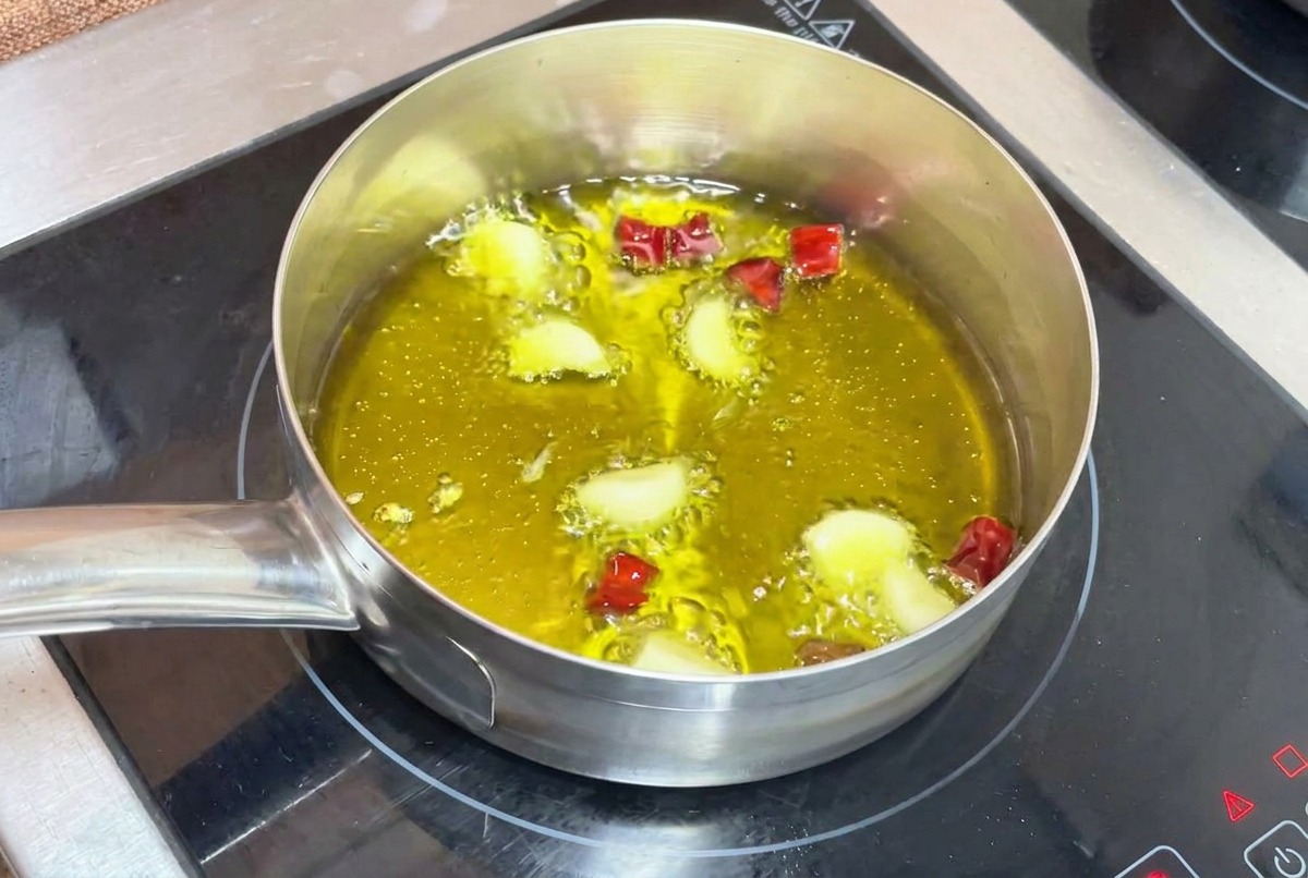 Garlic cloves and chopped dried chilies gently frying in bubbling hot olive oil inside a small metal pot on an induction stove.