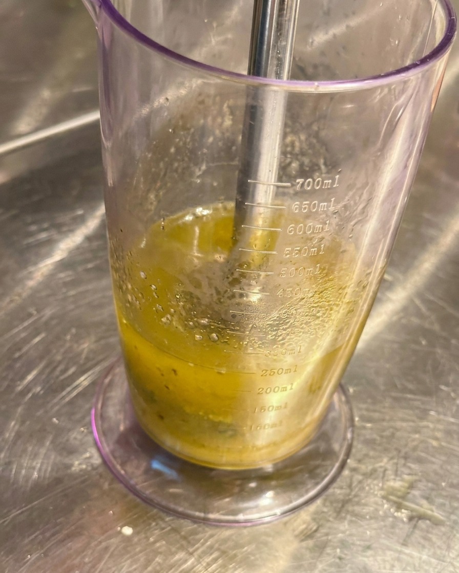 An immersion blender submerged in a tall clear plastic measuring beaker, blending roasted garlic and olive oil into a pale liquid paste.