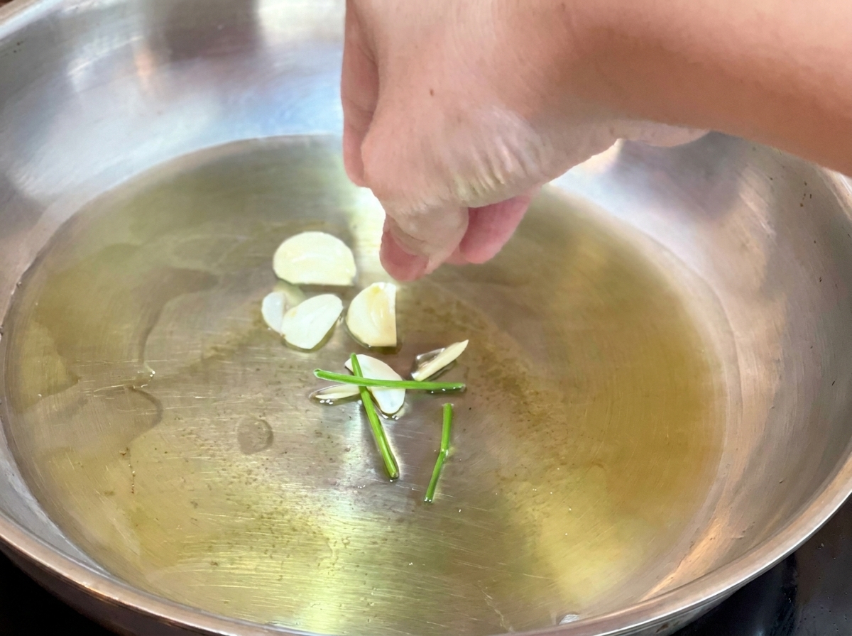 A hand dropping raw garlic slices and green parsley stems into hot olive oil in a frying pan.
