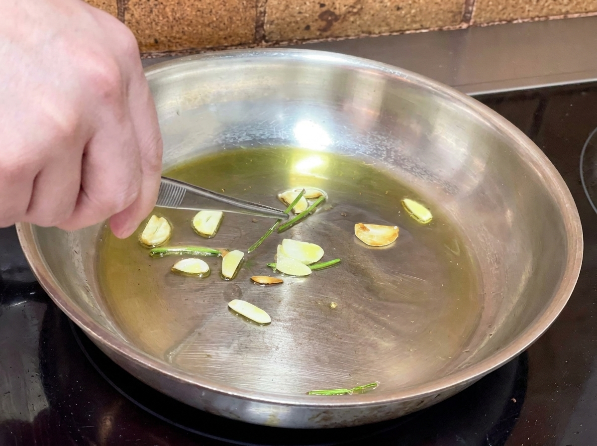 Metal tweezers picking out fried parsley stems from a pan of garlic-infused olive oil.