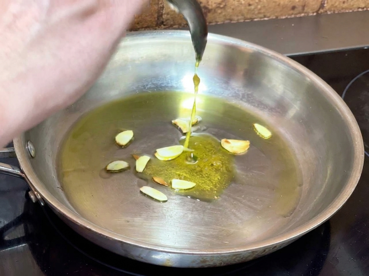 Spooning prepared garlic paste into a frying pan with infused olive oil and sliced garlic.