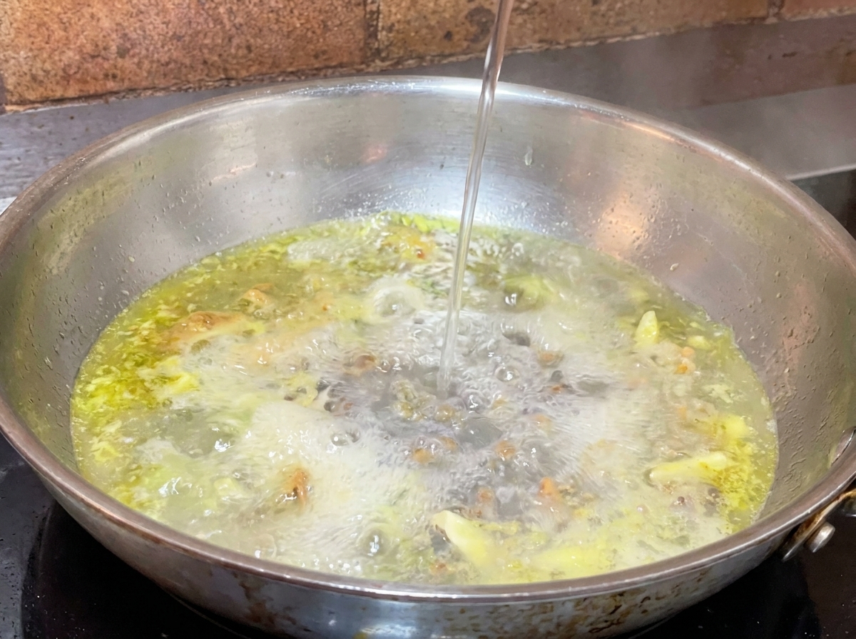 A thin stream of pasta water being poured into a hot pan of bubbling oil and garlic.